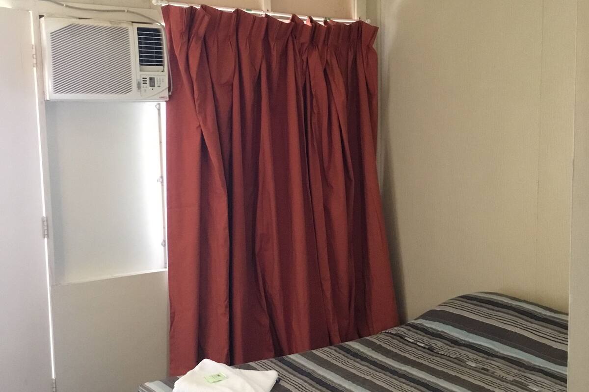 Una cama individual con ropa de cama a rayas, una cortina roja y un equipo de aire acondicionado.