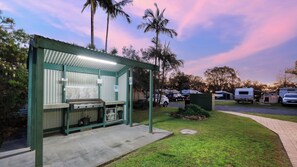 A covered outdoor kitchen area with a grill, surrounded by a grassy lawn and palm trees. - Anchorage Holiday Park - Iluka (Iluka)