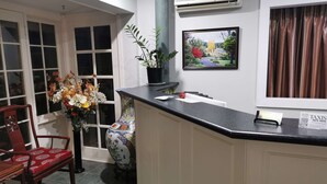 Un área de recepción con mostrador, una silla, un jarrón de flores, una planta en maceta, un cuadro enmarcado y un equipo de aire acondicionado.