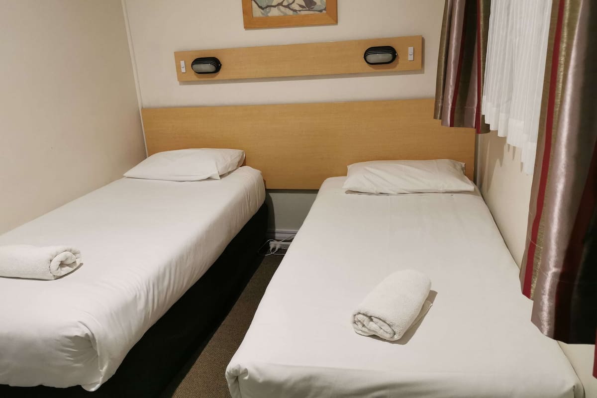 A hotel room with two single beds, each with a white bedspread and a rolled white towel. There is a wooden headboard, a small framed artwork on the wall, and a window with curtains.