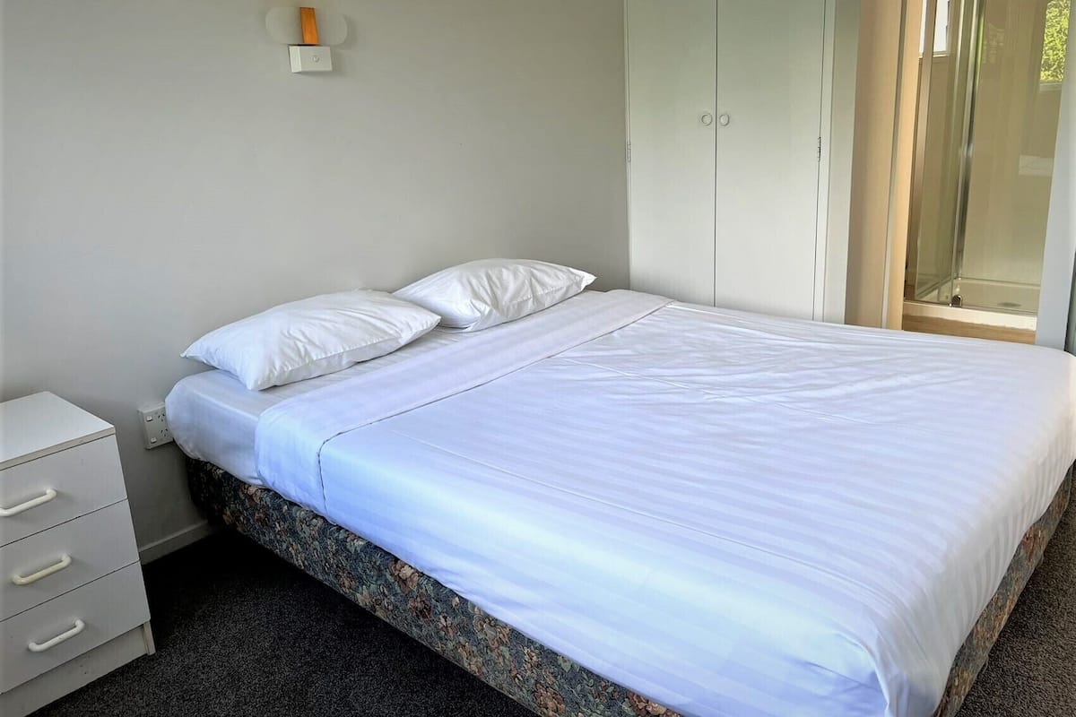 A neatly made bed with white linens, a small bedside table with drawers, and a wardrobe with a mirror.