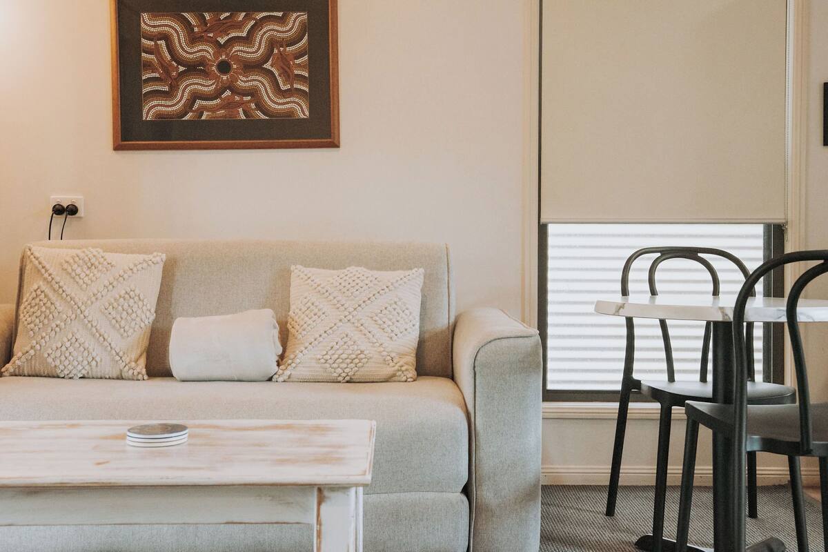 A living room with a sofa, two patterned pillows, a small table with a plate, a framed artwork on the wall, and a window with blinds.