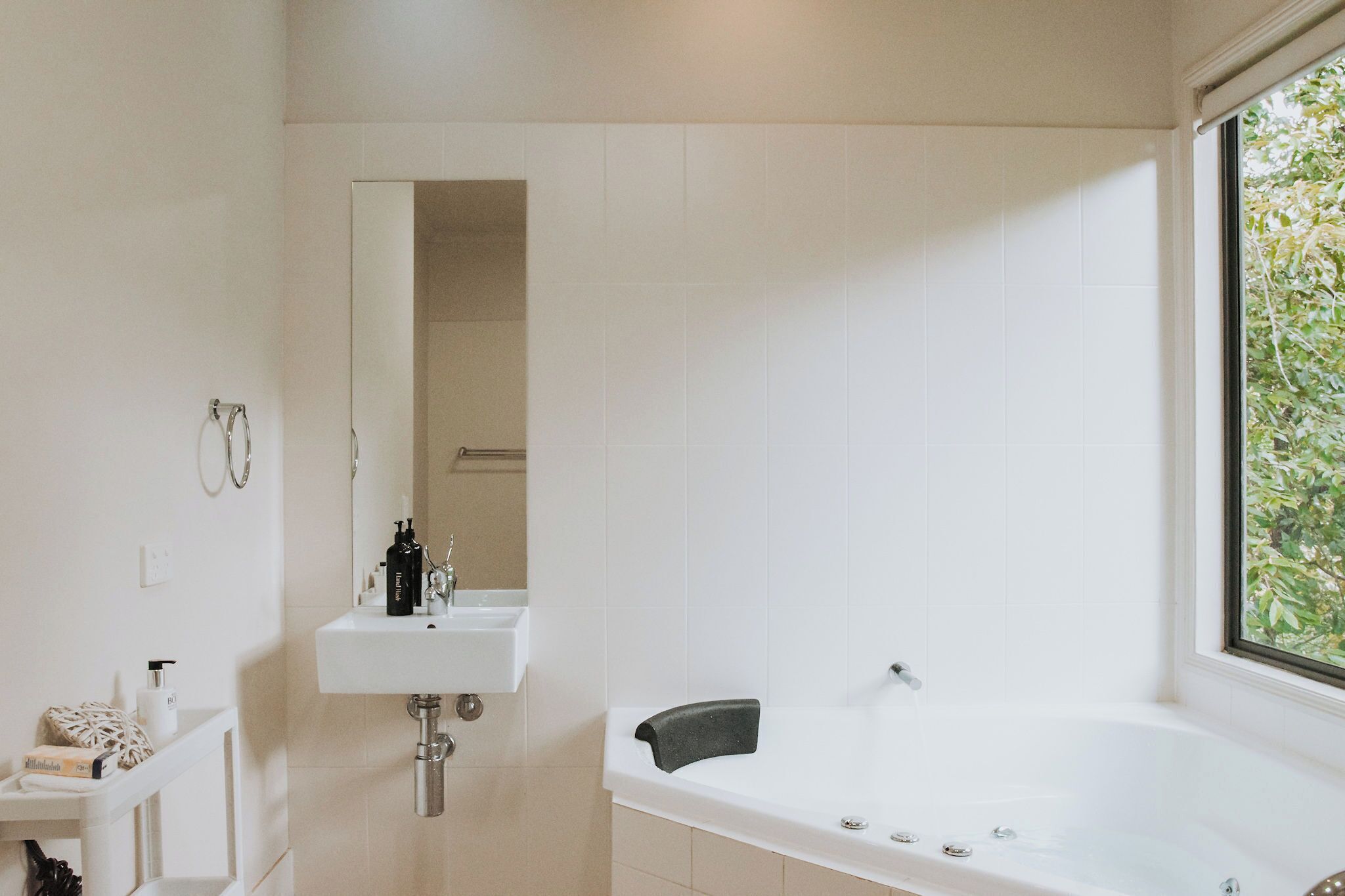 A modern bathroom with a white sink, a mirror, a bathtub, and a window with a view of greenery.