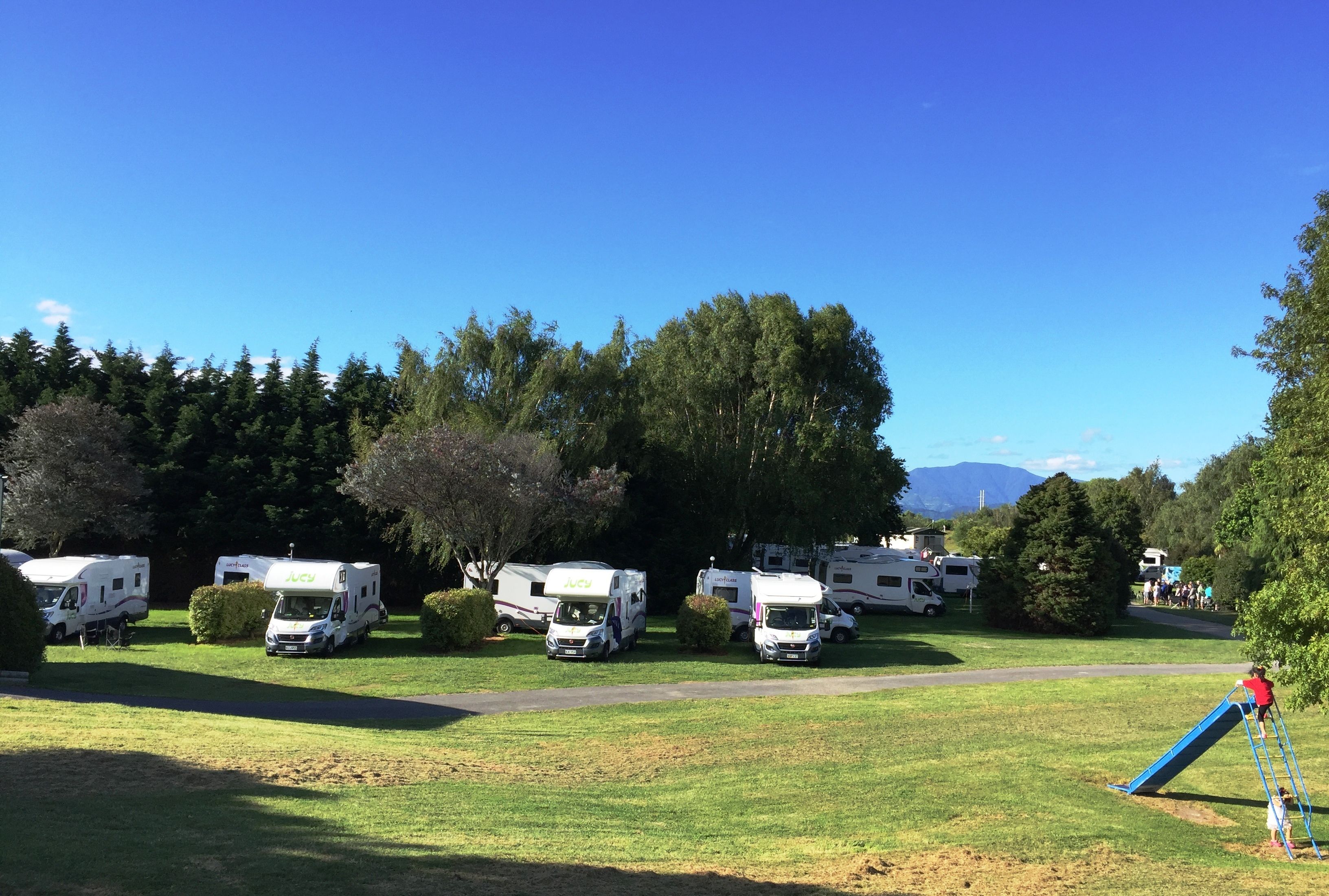 Un terrain de camping où des camping-cars sont stationnés sur une pelouse, entouré d’arbres et d’une aire de jeux.