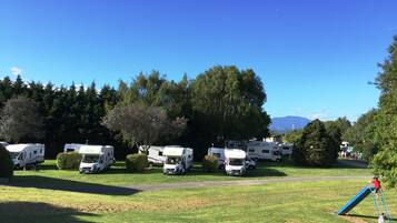 Un terrain de camping où des camping-cars sont stationnés sur une pelouse, entouré d’arbres et d’une aire de jeux.