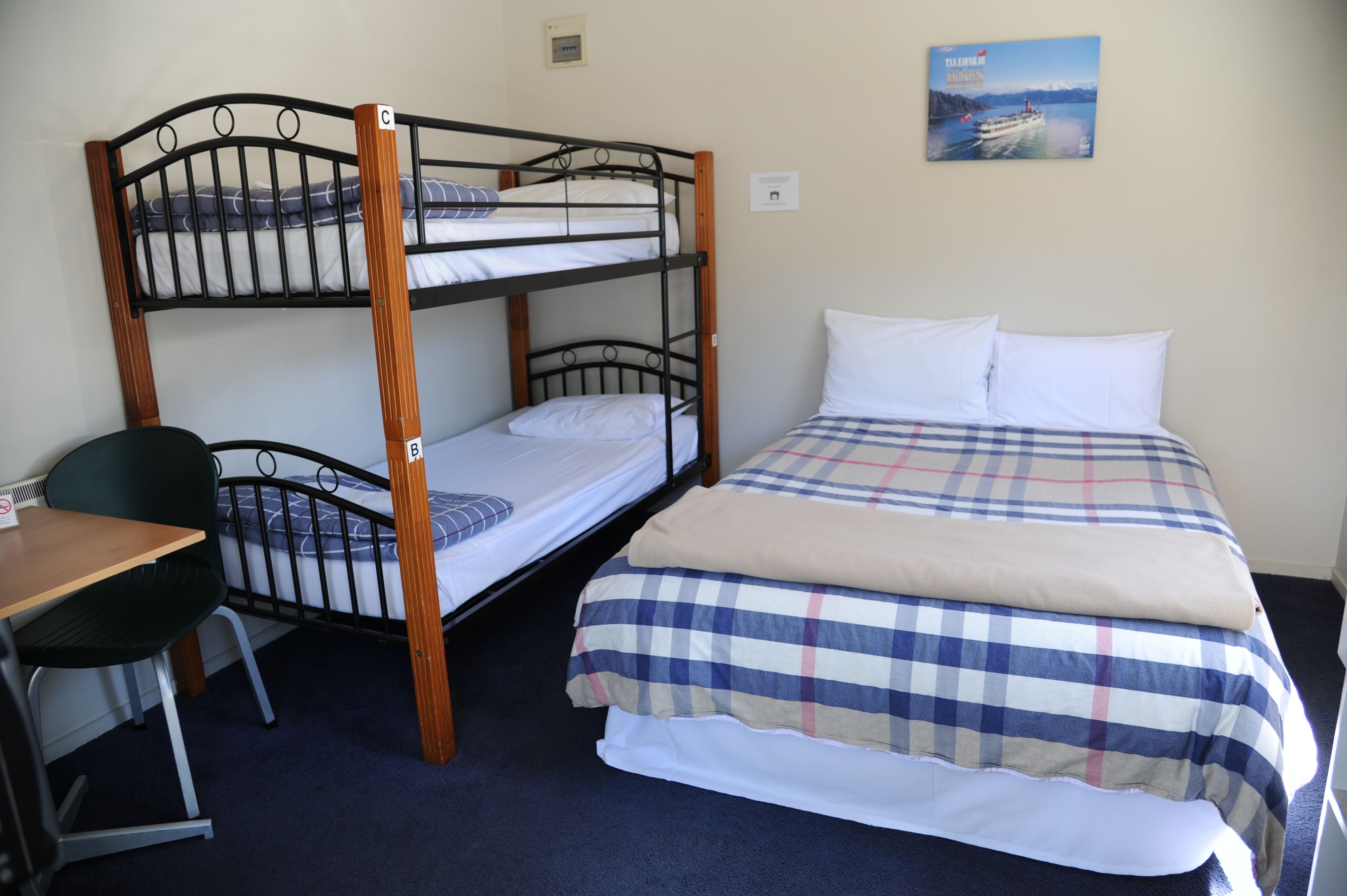 Uma cama beliche com escrivaninha e cadeira, uma cama de solteiro com colcha xadrez, e um quadro pendurado na parede de um barco.