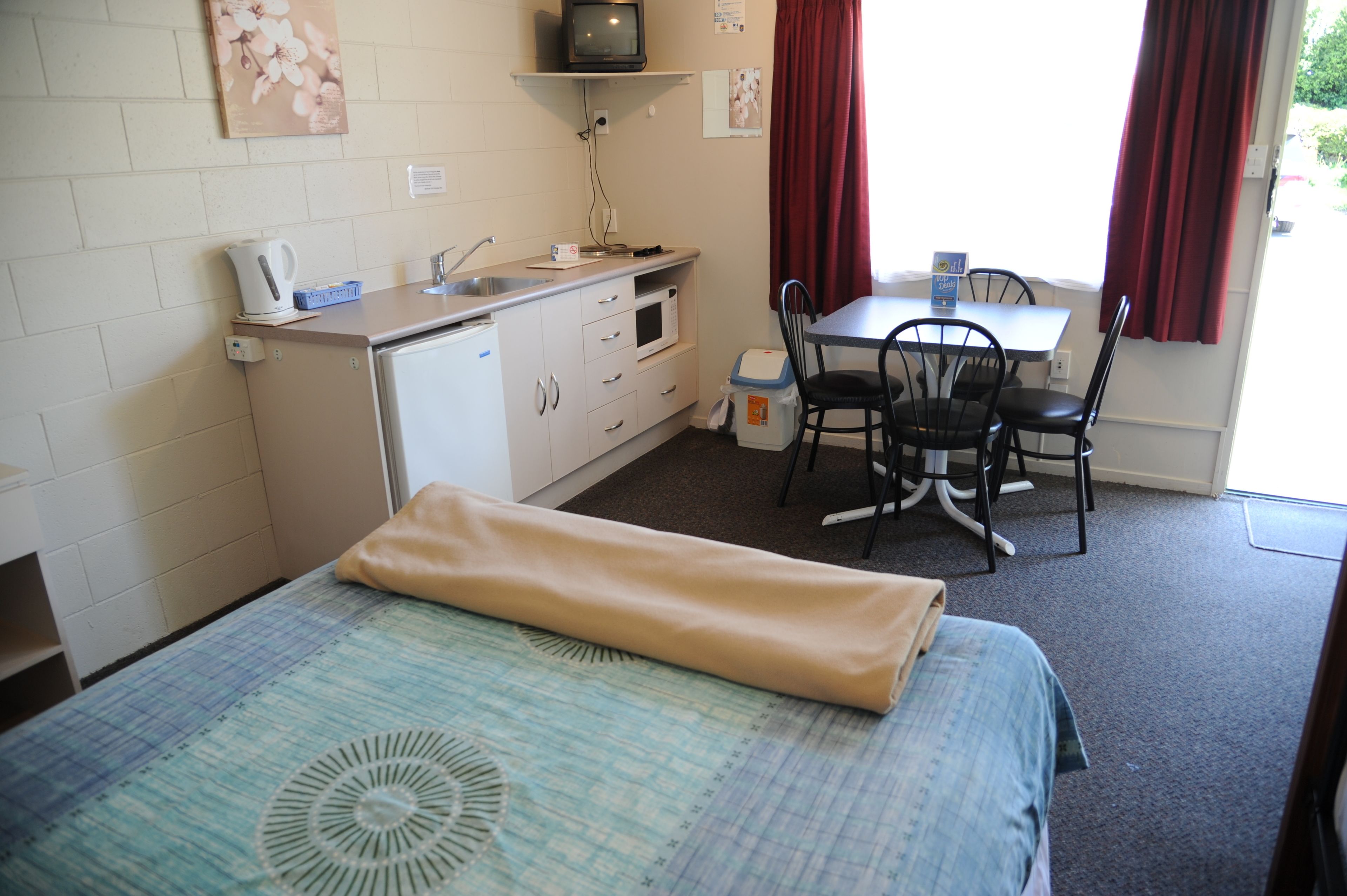 A small, compact room with a bed, a kitchenette, a dining table with chairs, and a window with curtains.