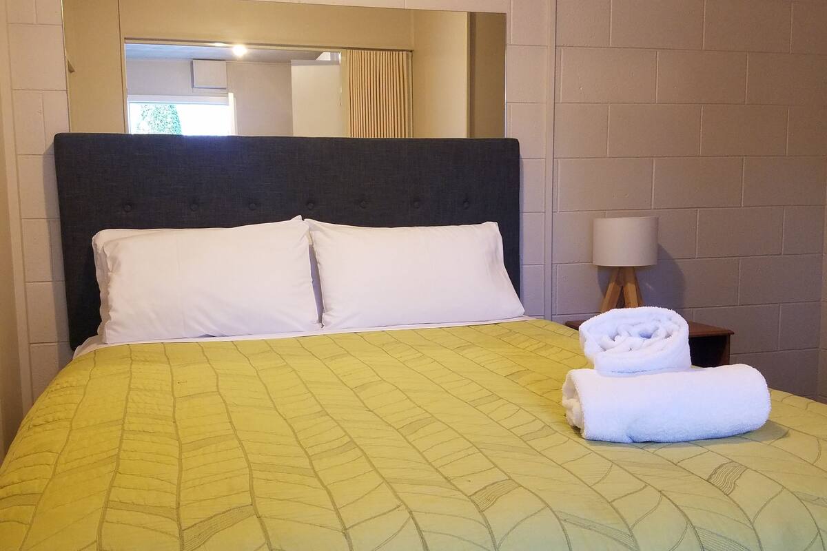 Ein Hotelzimmer mit einem Bett, weißen Kissen, einer gelben Bettdecke, einem gefalteten weißen Handtuch, einem Nachttisch mit Lampe und einem Spiegel über dem Bett.