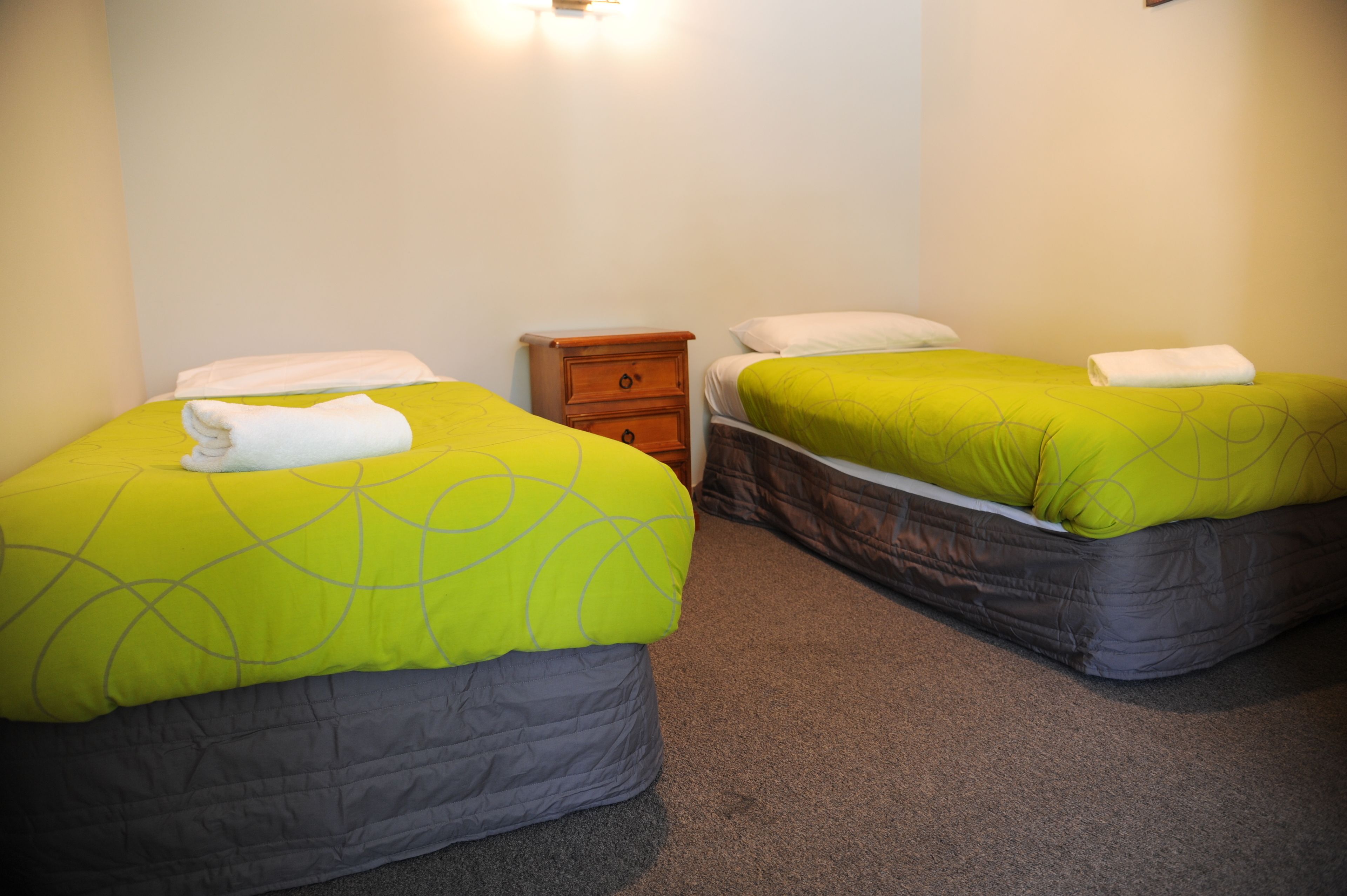 A room with two beds, each with a green bedspread and white pillows. There is a wooden nightstand between the beds.