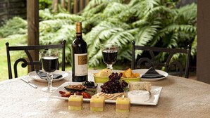 A table set with a bottle of wine, glasses, cheese, crackers, and grapes, with a backdrop of ferns. - B&B @ the Redwoods (Rotorua)