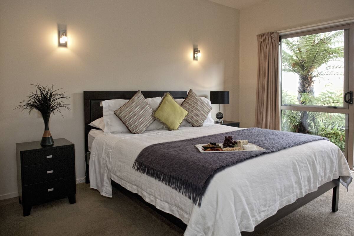 A neatly made bed with a white comforter and multiple pillows, a bedside table with a lamp, and a window with curtains.