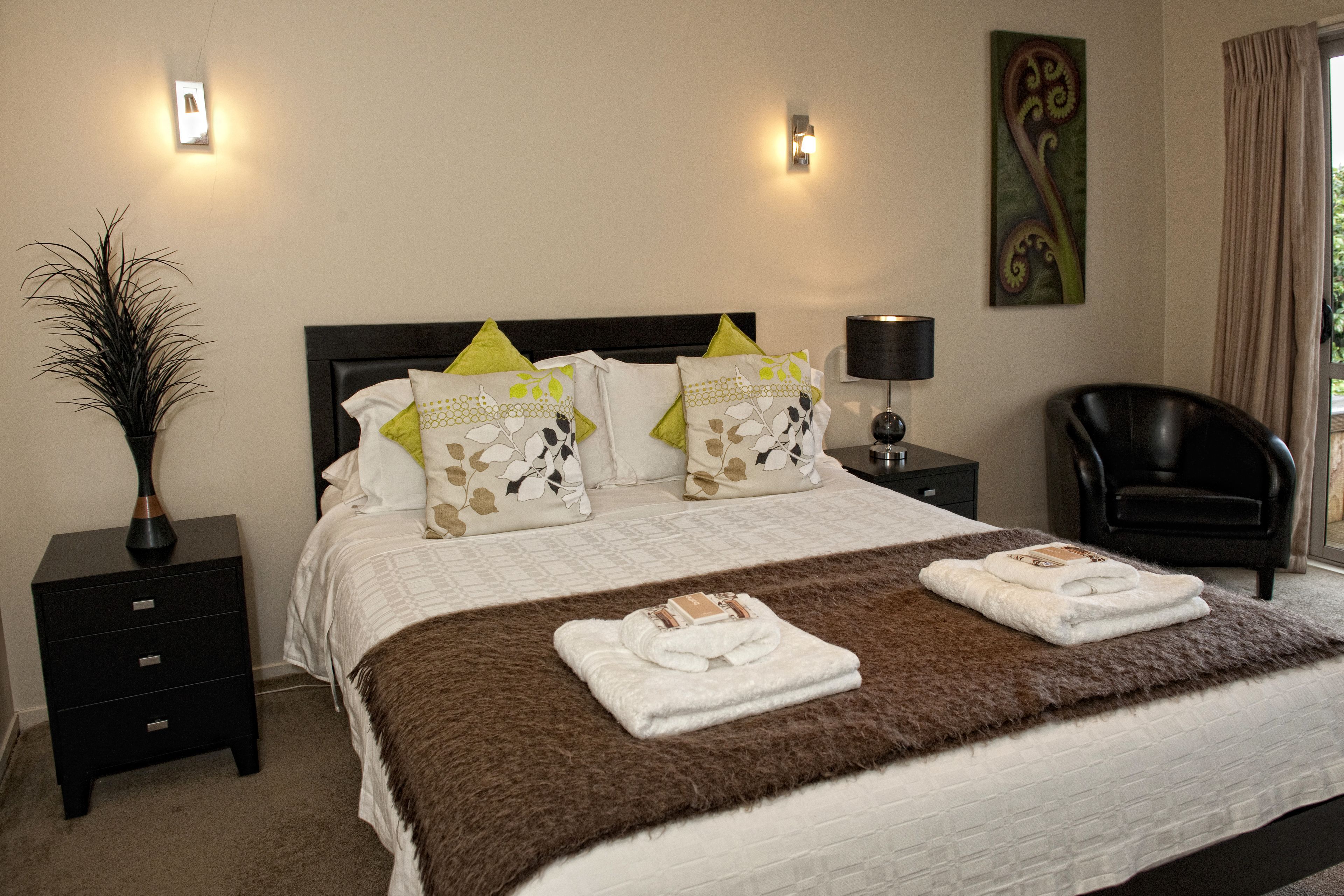 A neatly made bed with a brown blanket and two folded towels, a black nightstand with a vase, and a chair in a hotel room.