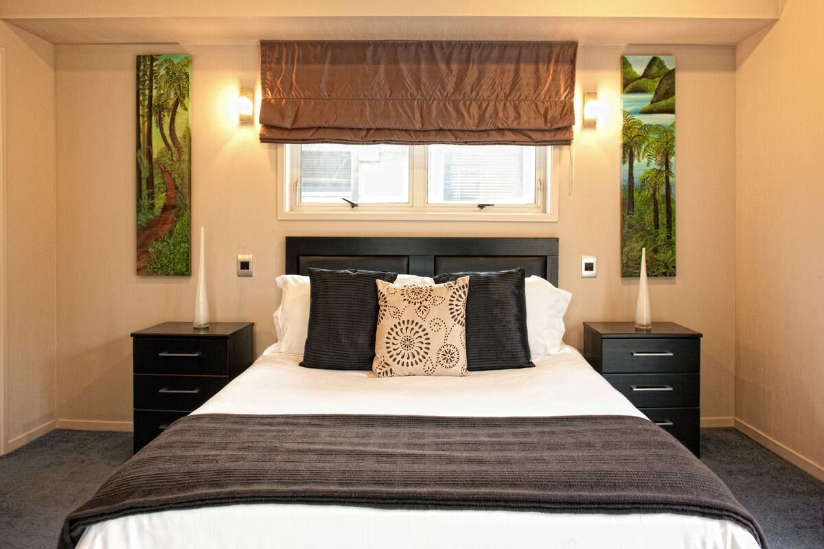A bedroom with a large bed, two bedside tables, a window with blinds, and two wall-mounted lamps.