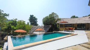 A rectangular swimming pool with a tiled deck, surrounded by trees and a thatched-roof structure.