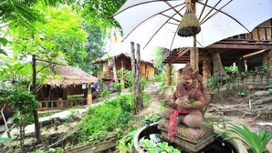 A rustic outdoor setting with a statue, thatched-roof structures, and lush greenery.