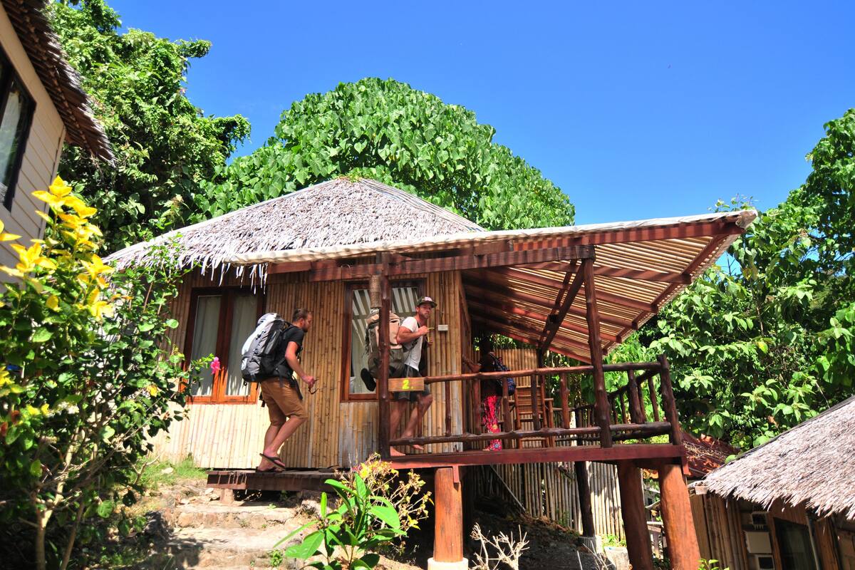 A wooden structure with a thatched roof, a wooden staircase leading to a porch, and a natural, green backdrop.