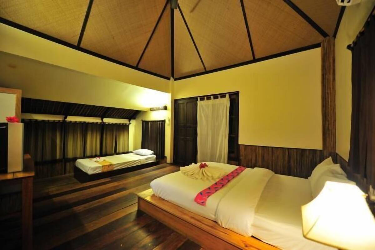 A hotel room with a wooden floor, a bed with white bedding and a red accent, a nightstand, a desk with a lamp, and a ceiling fan.