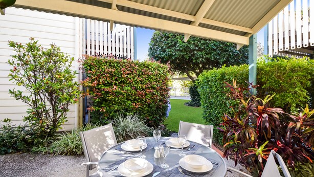 An outdoor patio with a round table set for four, surrounded by chairs, and a covered area with a white fence. - Terrapin Apartments (Noosaville)