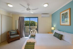 Une chambre d’hôtel avec un lit, une chaise en osier, un balcon avec vue et un ventilateur de plafond.