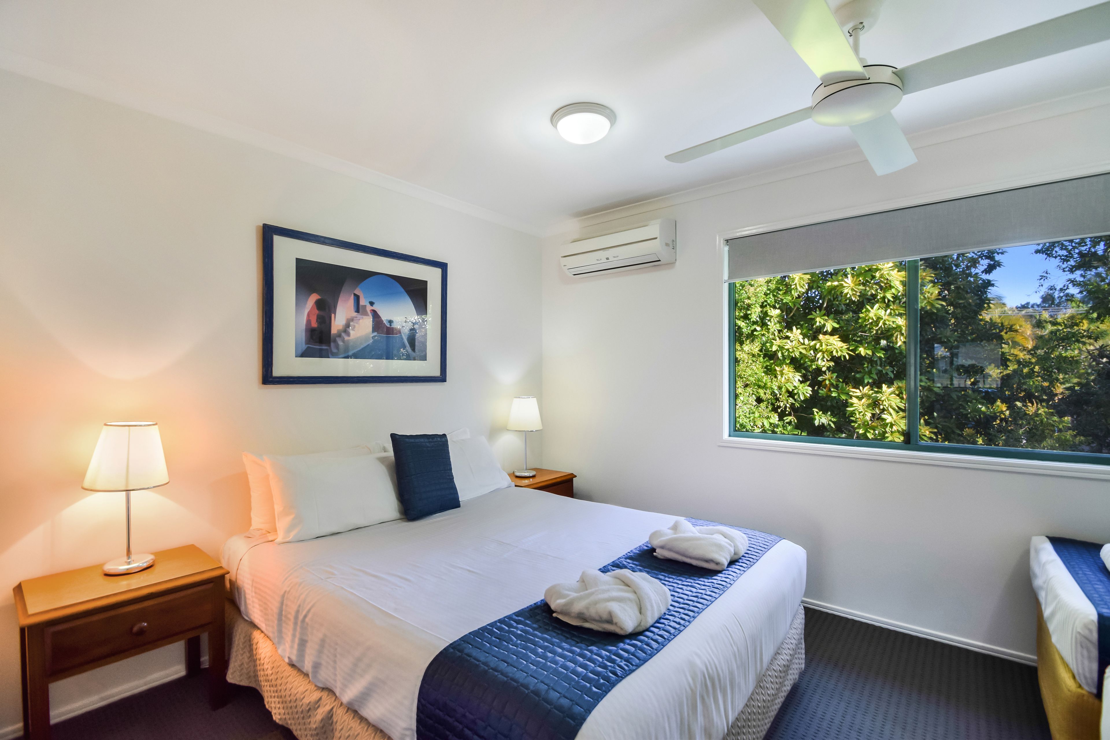 Habitación de hotel con cama, mesita de noche, lámpara, ventilador de techo, aire acondicionado y ventana con vistas a árboles.