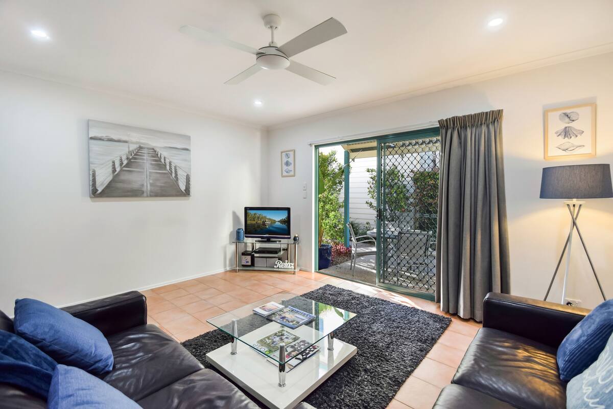 A modern living room with a glass coffee table, black leather sofas, a ceiling fan, and a TV on a stand.