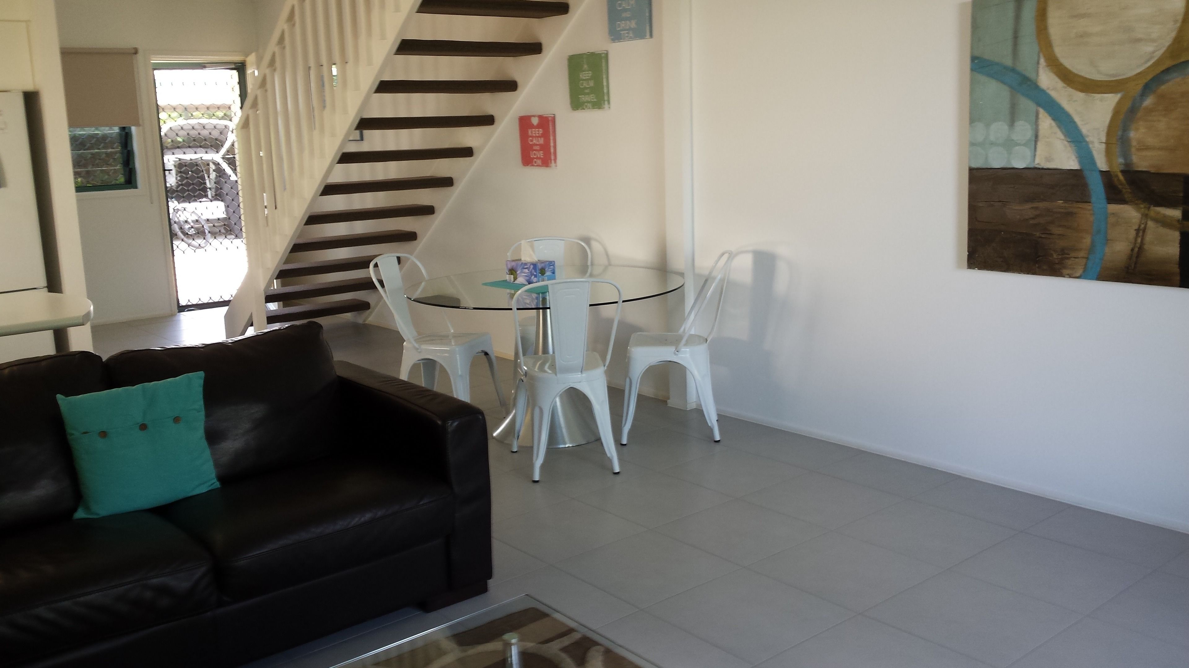 A modern living room with a black leather sofa, a glass coffee table, and a dining area with white chairs and a glass table. A staircase with wooden steps is visible in the background.