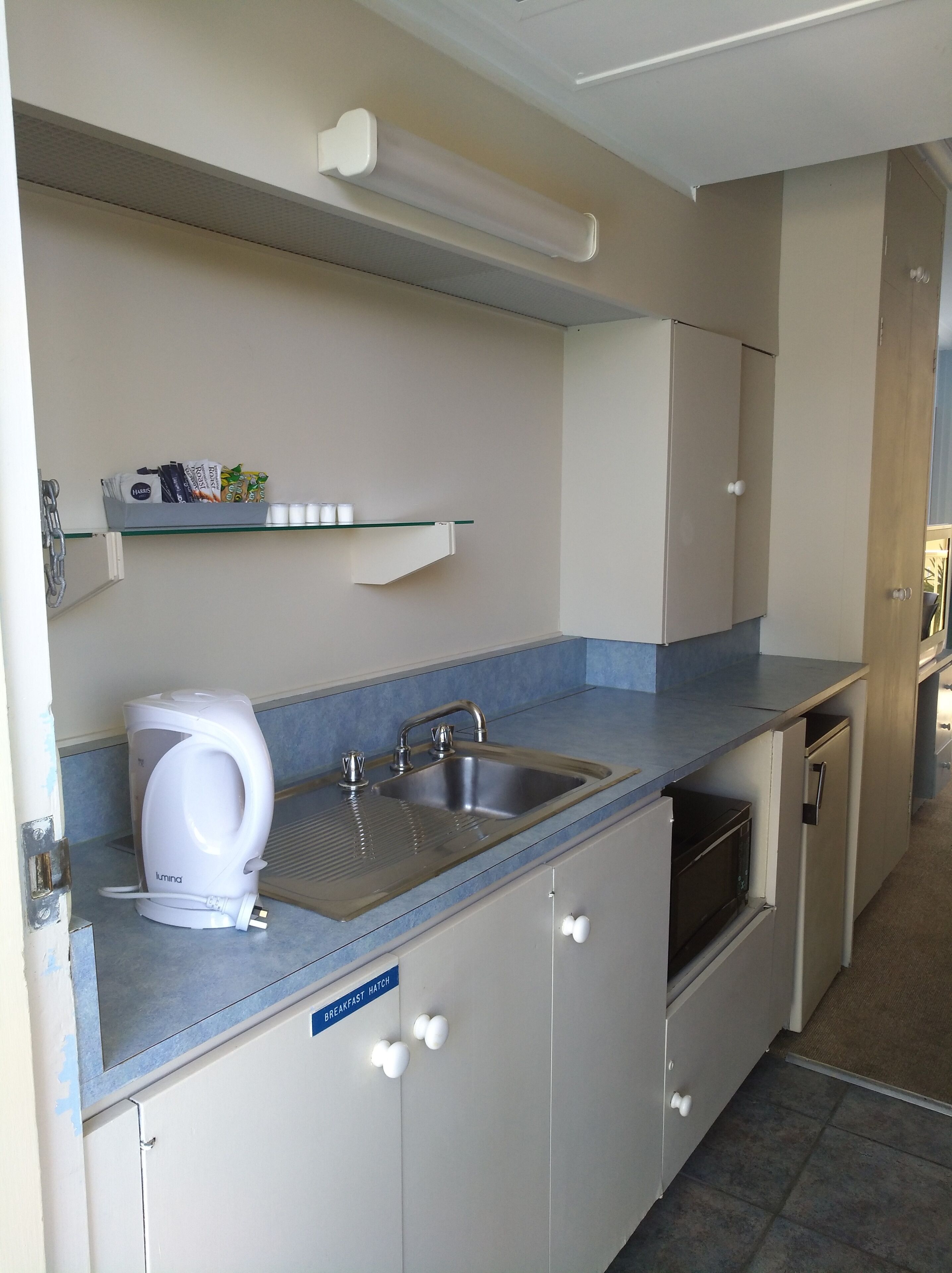 A compact kitchen with a built-in sink, a kettle, and a microwave. There is a shelf above the sink with various items.