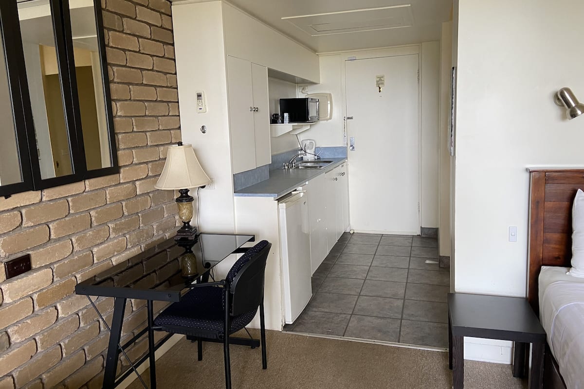 A compact hotel room with a kitchenette, a dining area with a chair and table, and a bed with a wooden headboard.