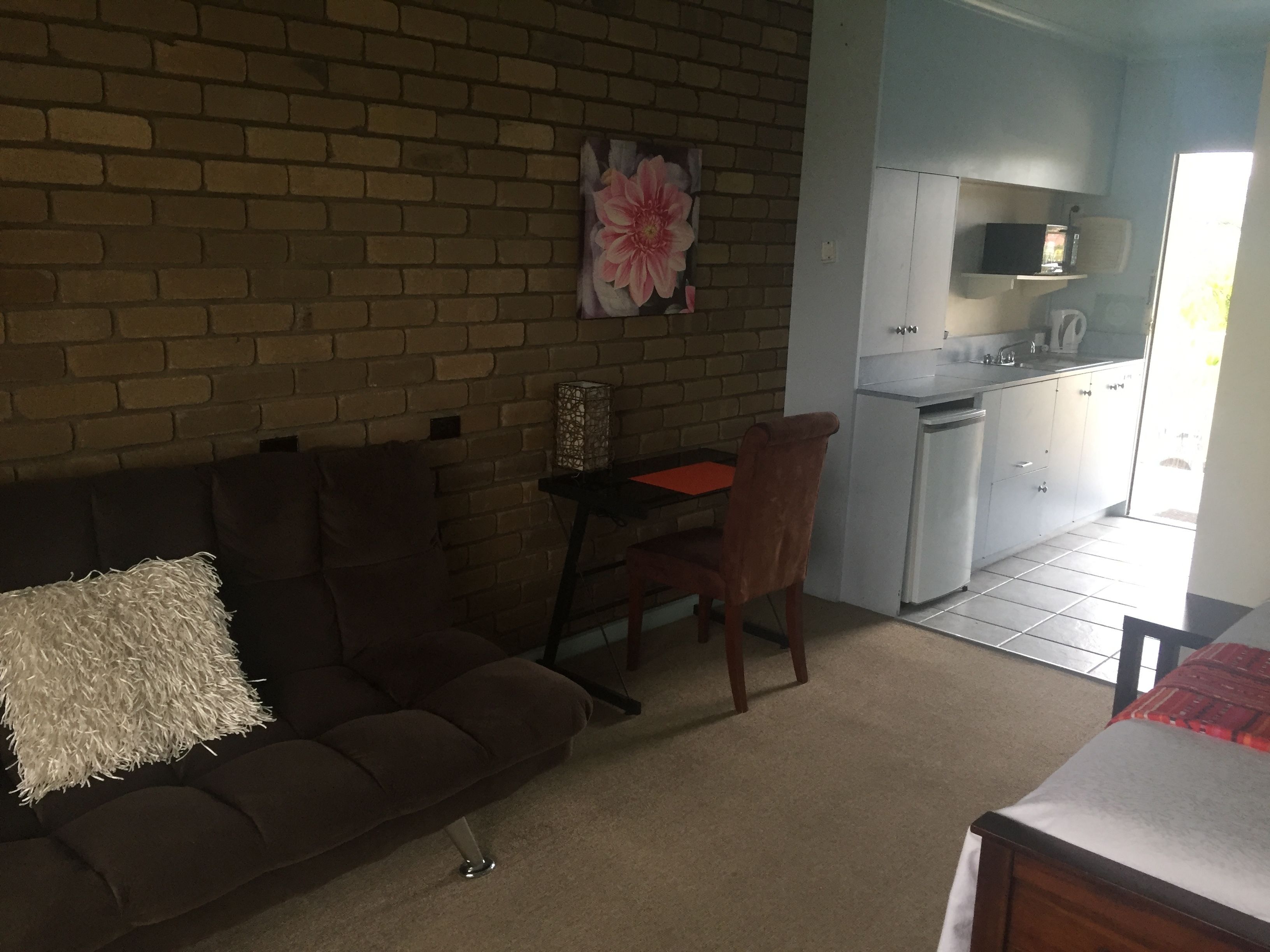 Una sala de estar con un sofá marrón, una mesita y una silla. Cuenta con una zona de cocina con microondas y armarios.