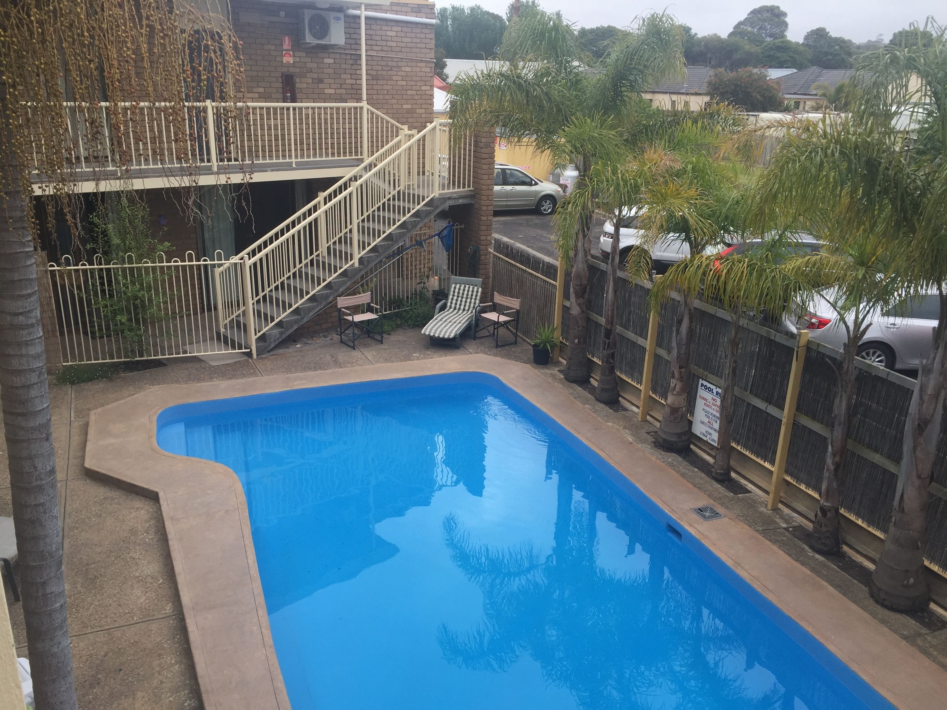 A rectangular swimming pool with a white deck and a small staircase leading to a balcony. There are two lounge chairs and a potted plant near the pool.