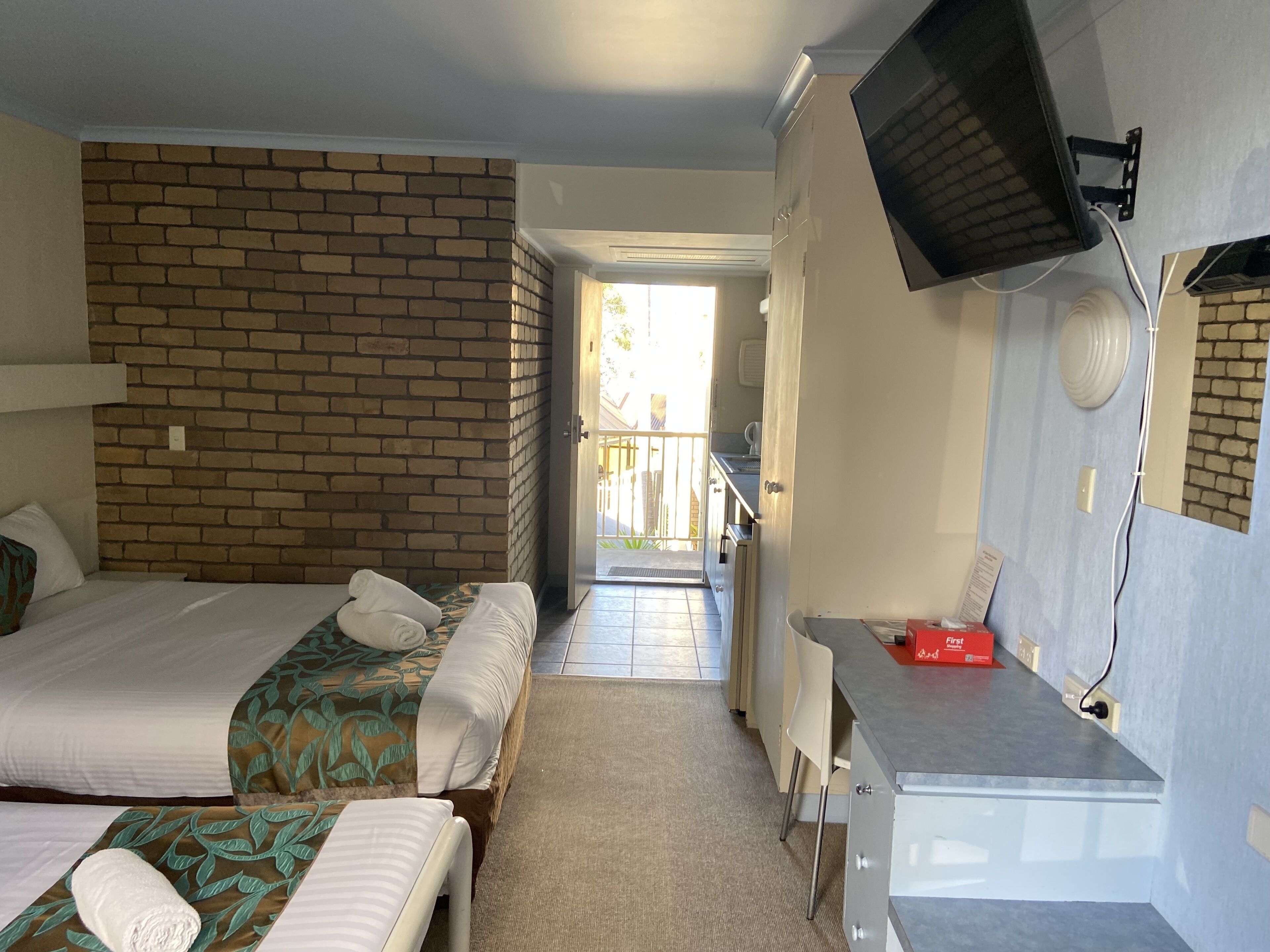 A hotel room with two beds, a desk, a TV, and a brick wall.