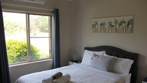 Ein ordentlich bezogenes Bett, gerollte Handtücher, ein Nachttisch mit Lampe, ein Fenster mit Vorhängen und ein Blumenbild an der Wand.