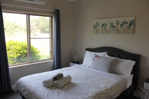 A neatly made bed with rolled towels, a nightstand with a lamp, a window with curtains, and a floral artwork on the wall.