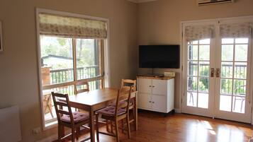 A dining area with a wooden table and chairs, a television on a cabinet, and large windows with curtains.
