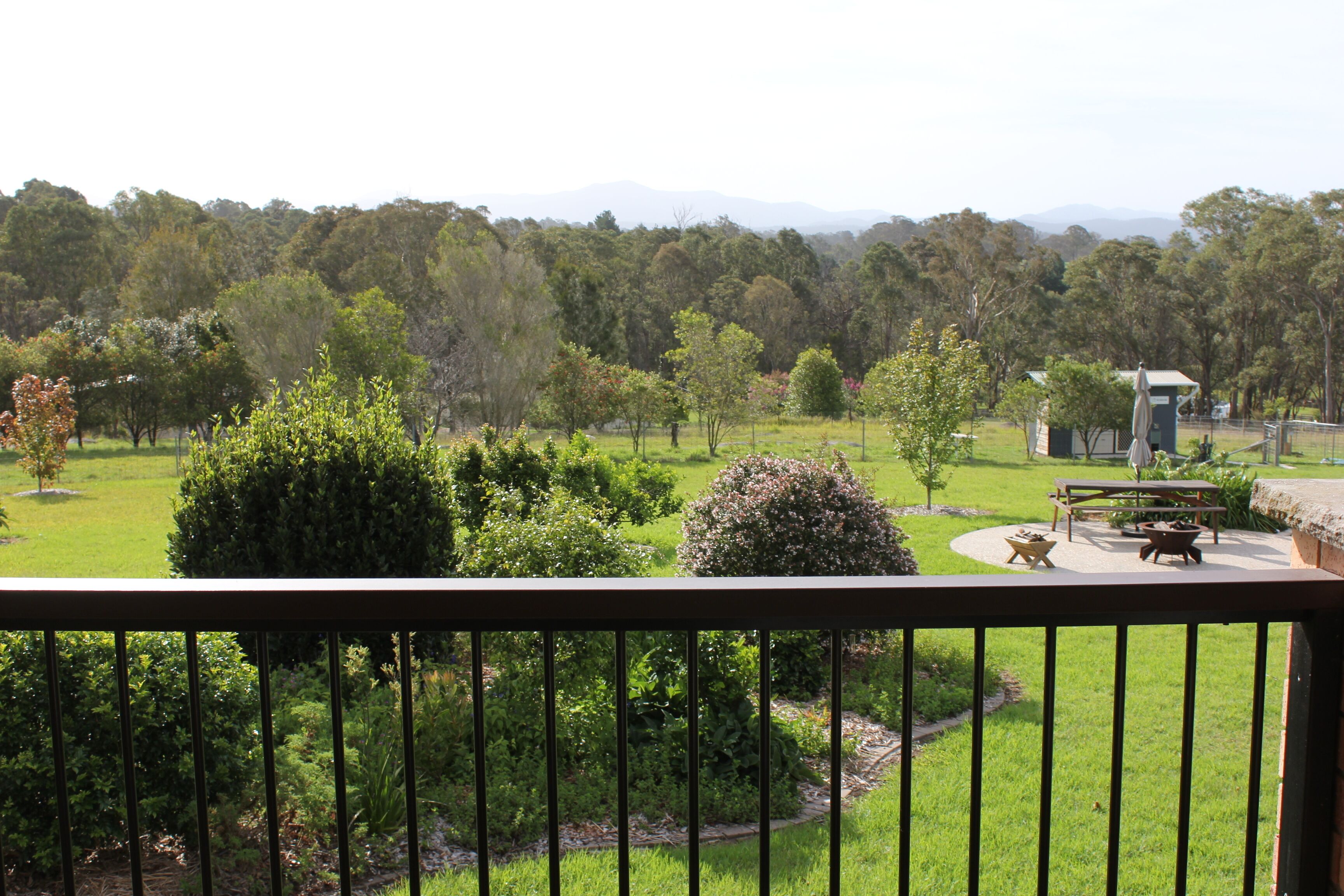 Vista desde un balcón sobre un jardín ajardinado con zona de picnic, un edificio pequeño y montañas al fondo.