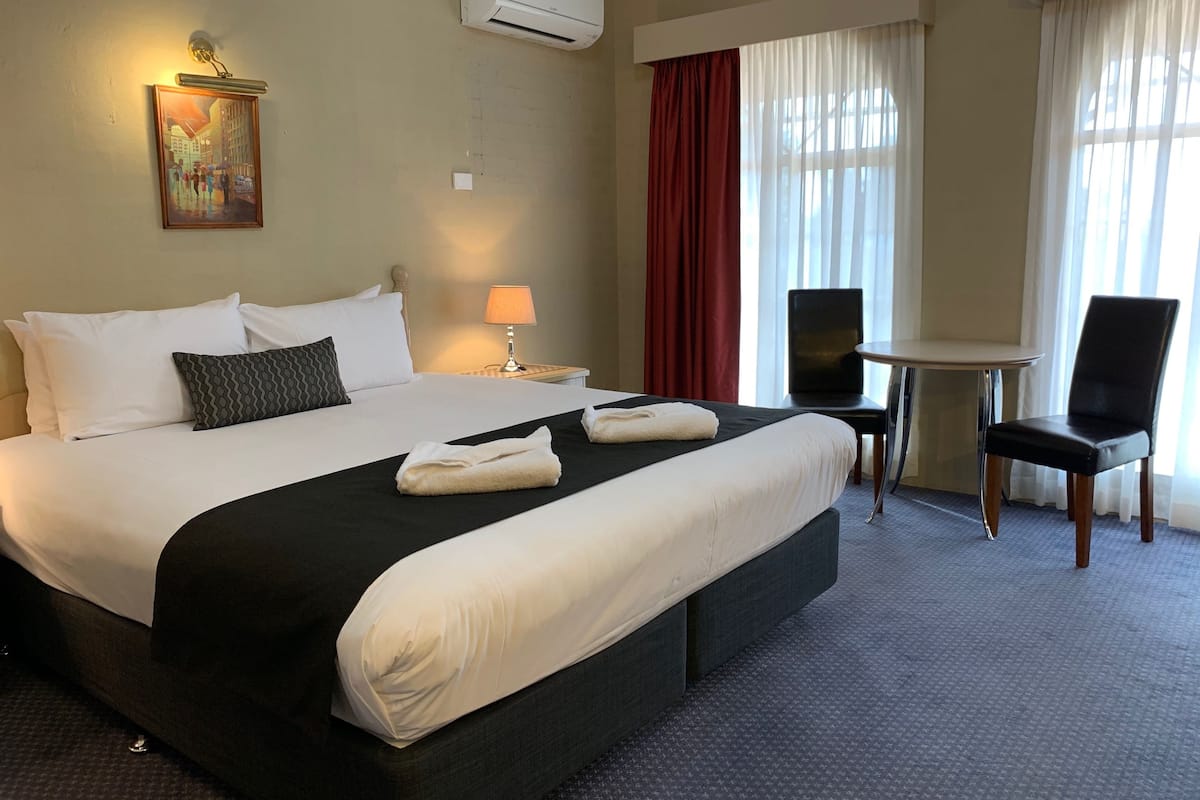 A hotel room with a large bed, a desk, a chair, an air conditioner, and a painting on the wall.