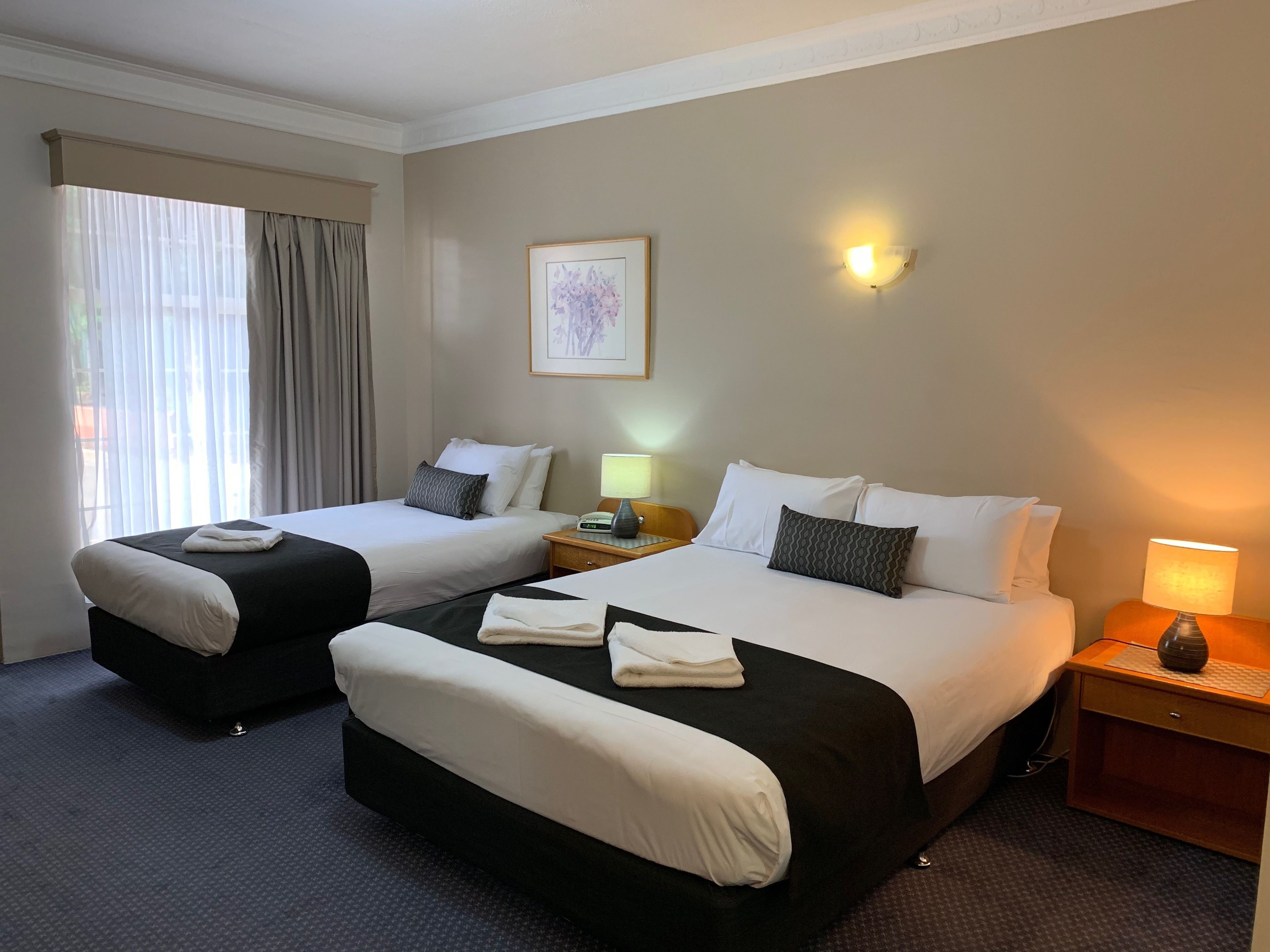 A hotel room with two beds, a bedside table with a lamp, a framed picture on the wall, and a window with curtains.