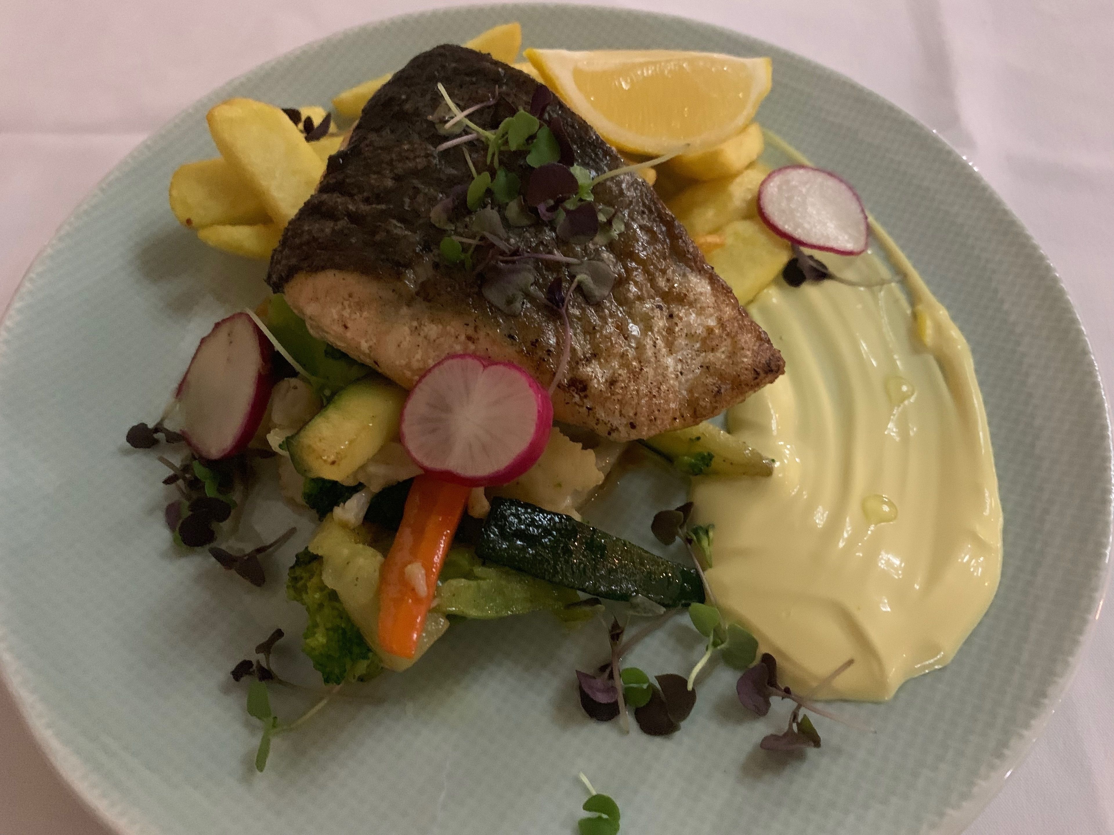 Une assiette contenant un morceau de poisson grillé, des tranches de citron, des radis et une sauce crémeuse.