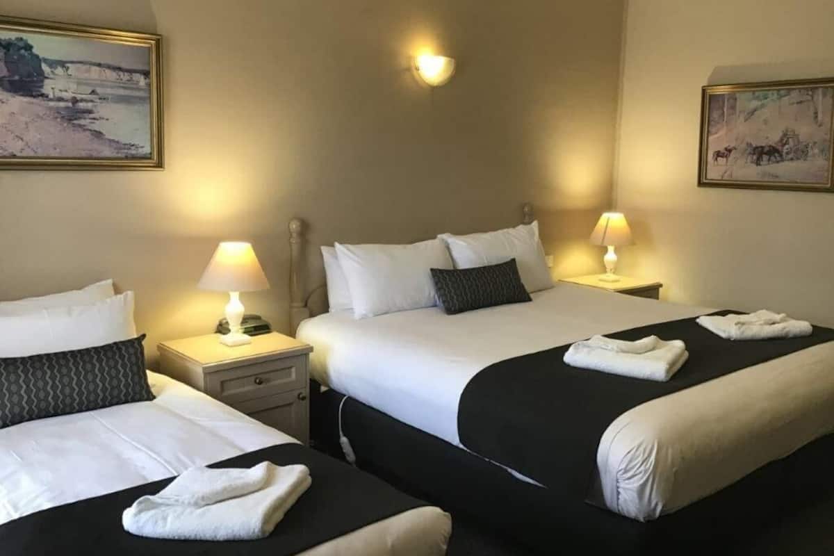 A hotel room with two beds, each with a black and white bedspread, white pillows, and a bedside lamp. There are paintings on the walls and a small table with a lamp.