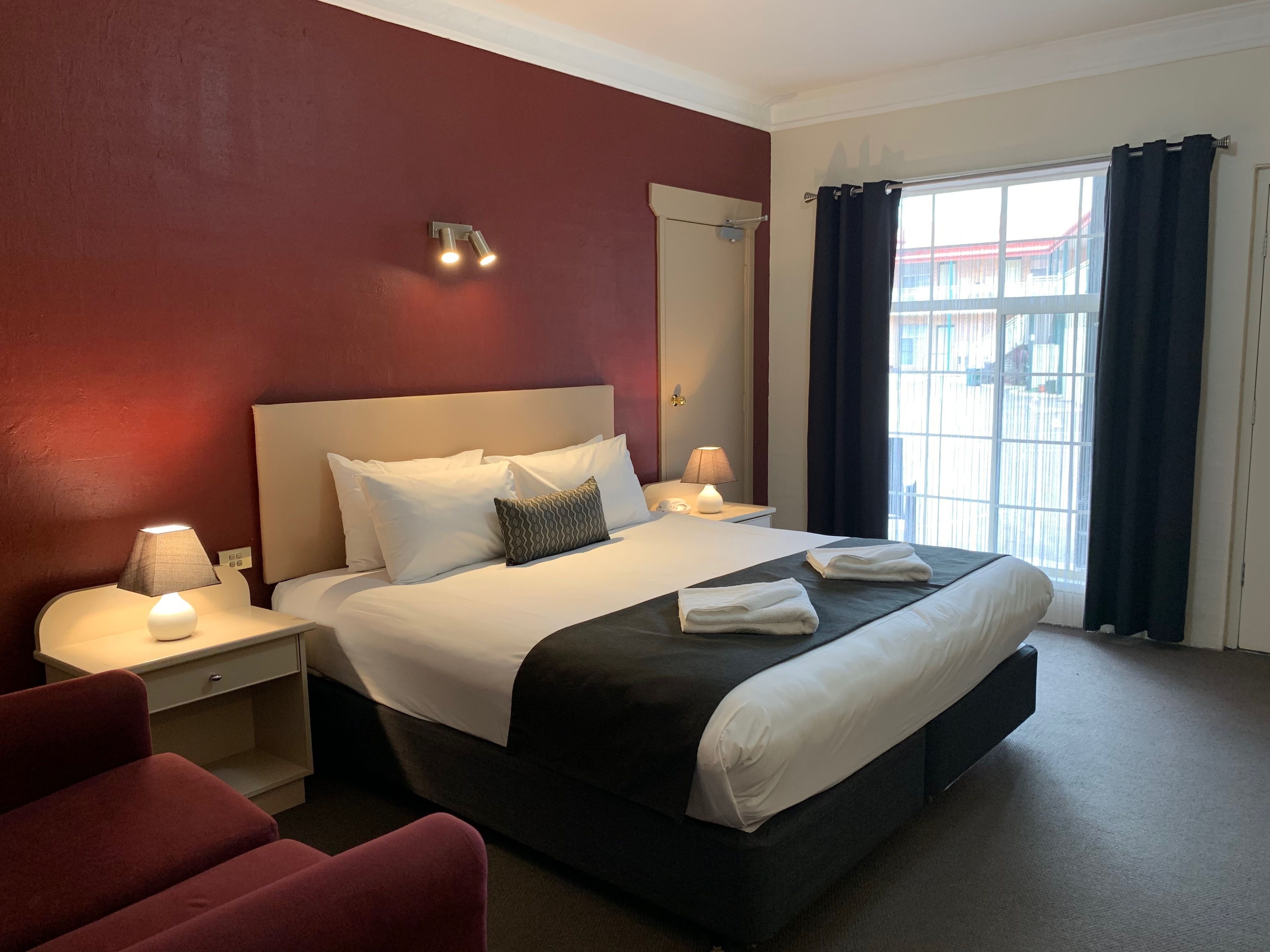 A hotel room with a large bed, two bedside tables with lamps, a red wall, a window with curtains, and a view of buildings outside.