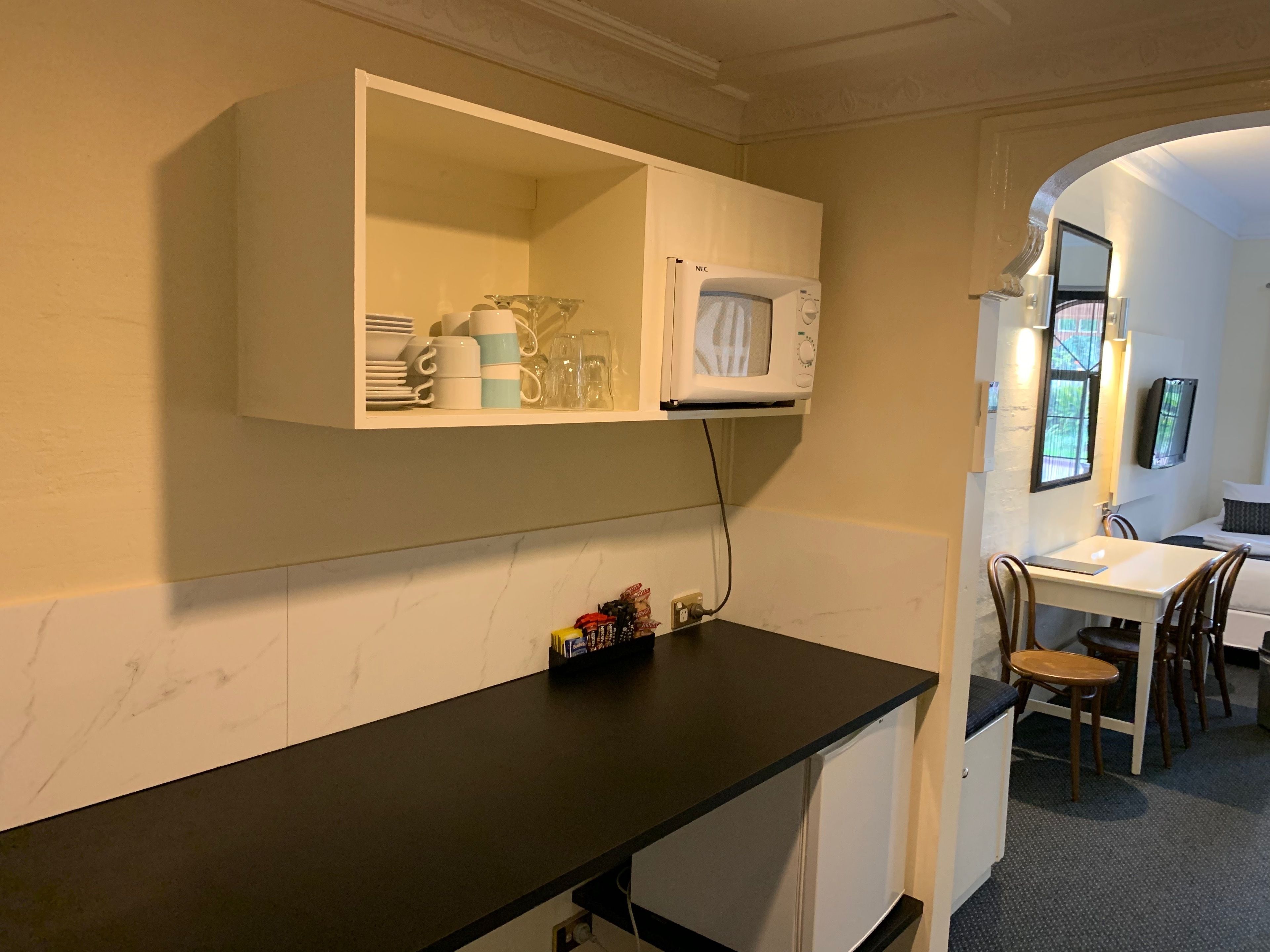 Een kitchenette met een magnetron, een plank met servies en een klein kastje.