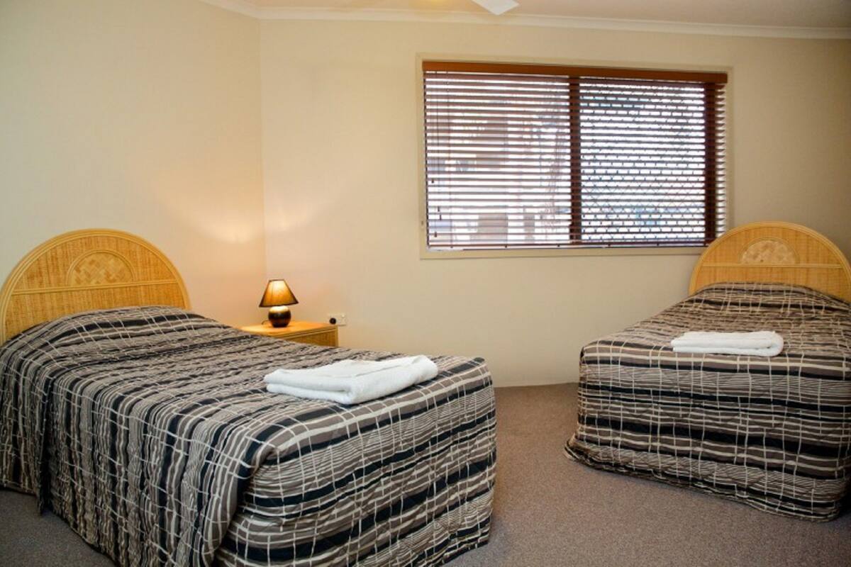 A room with two beds, each with a headboard, a bedside table with a lamp, and a window with blinds.