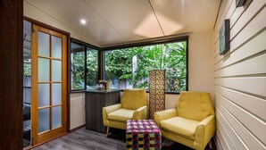 Una habitación con dos sillones amarillos, un reposapiés a cuadros, una puerta de madera y vistas a zonas verdes a través de amplios ventanales.