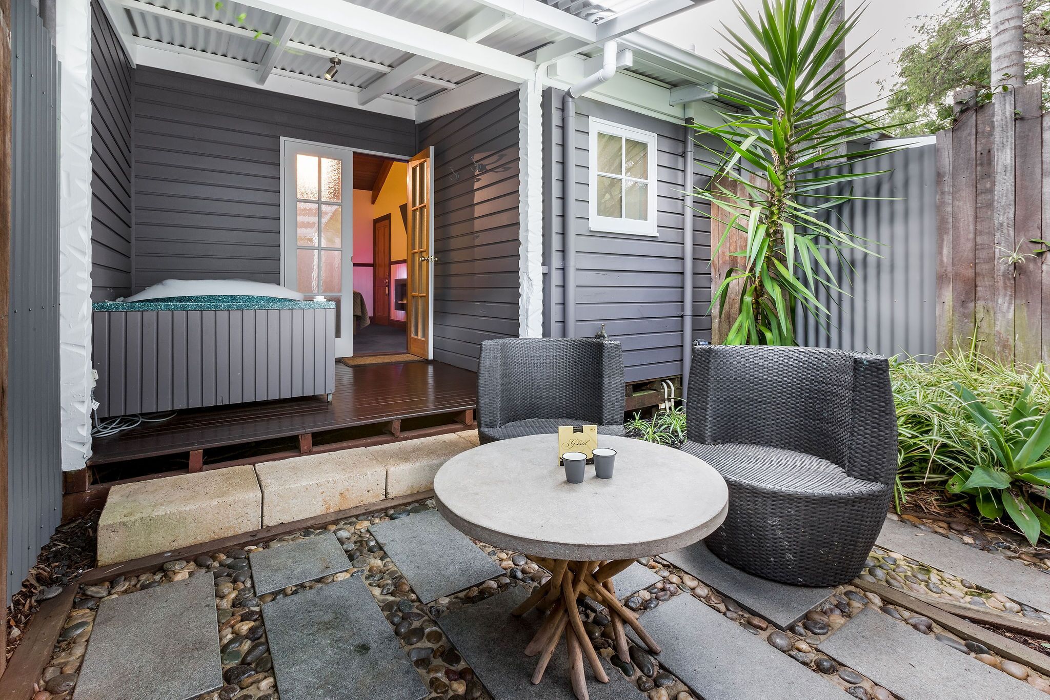 Un espace extérieur accueillant avec une table ronde et deux chaises, un petit lit avec une tête de lit verte et une clôture en bois agrémentée de plantes.