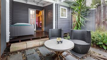 Un espace extérieur accueillant avec une table ronde et deux chaises, un petit lit avec une tête de lit verte et une clôture en bois agrémentée de plantes.
