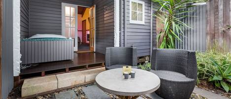 Een knus buitgedeelte met een ronde tafel en twee stoelen, een klein bed met een groen hoofdeinde, en een houten omheining met planten.