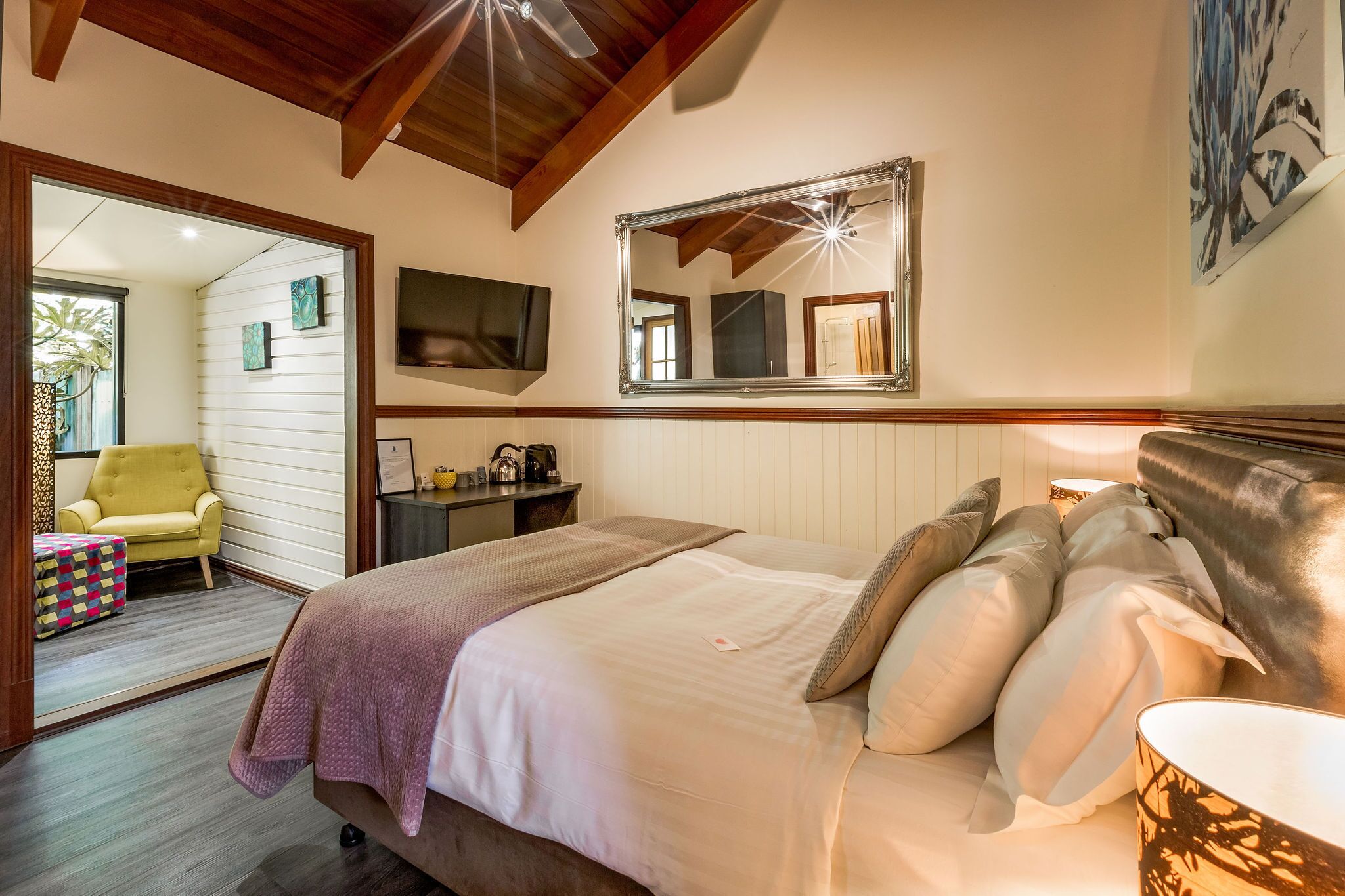 Un dormitorio con una cama grande, techo de madera, televisor, espejo y una mesita de noche con lámpara.