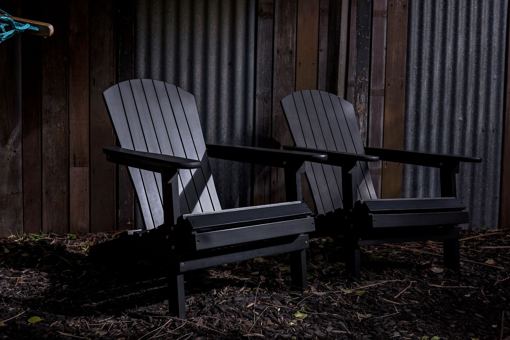 Dos sillas Adirondack con estructura metálica y listones de madera, ubicadas sobre una superficie de grava con una valla de madera al fondo.