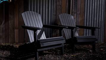 Two Adirondack chairs with metal frames and wooden slats, positioned on a gravel surface with a wooden fence in the background.