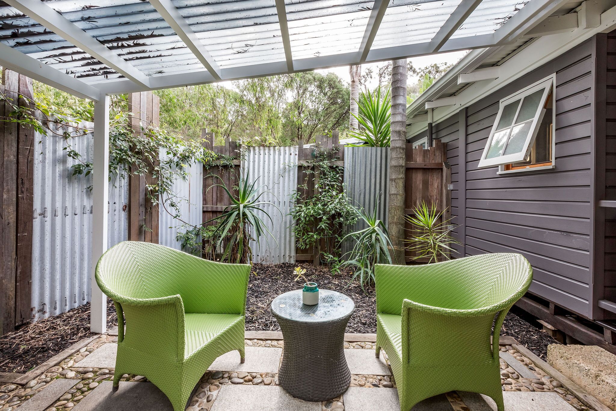 Überdachte Terrasse mit zwei grünen Korbstühlen und einem runden Tisch, umgeben von einem Holzzaun und verschiedenen Pflanzen.